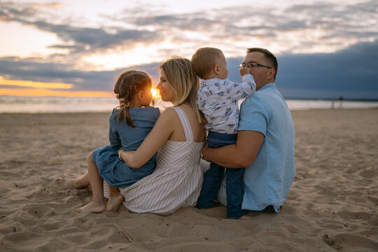 Happy family on the beach sitting on the sand on sunset,boy trying to take glasses of fathers face. Mom looking at daughter. Image with selective focus.