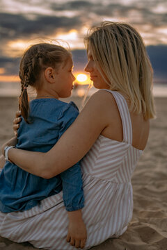 young blond caucasian woman sitting with her daughter on beach looking at sunset. Time together. Family concept. Image with selective focus