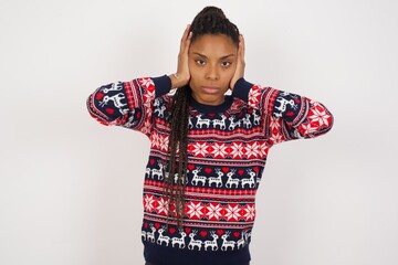 Frustrated Young beautiful African American woman wearing Christmas sweater against white wall plugging ears with hands does not wanting to listen hard rock, noise or loud music.