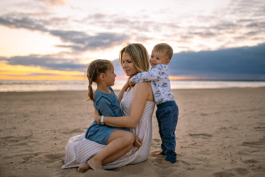 portrait of happy beautiful mother with her daughter sitting on her laps and baby son hugging her on the beach in sunset. Beautiful cloudy sky on background. Image with selective focus