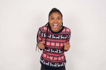 Young beautiful African American woman wearing Christmas sweater against white wall raising fists...