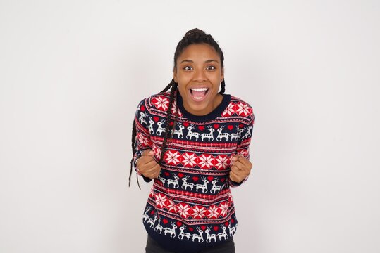 Young beautiful African American woman wearing Christmas sweater against white wall raising fists up screaming with joy being happy to achieve goals.