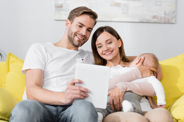 digital tablet in hand of man near happy wife holding in arms infant son