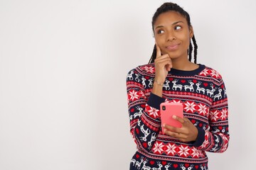 Thinking dreaming Young beautiful African American woman wearing Christmas sweater against white wall using mobile phone and holding hand on face. Taking decisions and social media concept.