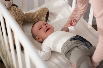 mother lifting crying infant son in baby crib on blurred foreground - obrazy, fototapety, plakaty