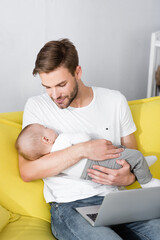 caring father holding in arms sleepy baby son while sitting on sofa with laptop
