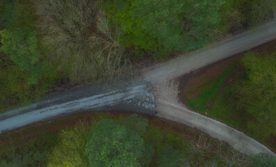 Y shaped crossroads visible from above. Green trees and grey road with transition from asphalt to gravel.
