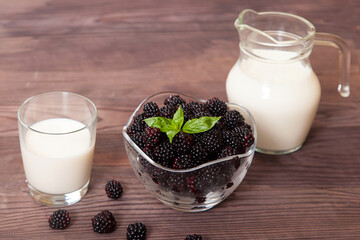 Blackberries in a glass cup, decorated with mint leaves, a glass of milk and a jug of milk on a wooden table. A healthy dessert.