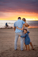 Happy family: blond mother, dad in blue shirt and jeans and little girl standing on the beach at sunset looking at the sea