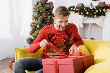Obraz na płótnie Canvas smiling man in red sweater holding ribbon on wrapped present while sitting on sofa with blurred christmas tree on background