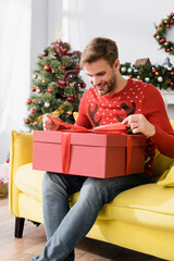 Obraz na płótnie Canvas happy man in red sweater looking at wrapped present while sitting on sofa with blurred christmas tree on background