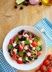 Traditional Greek Salad with English cucumbers,juicy tomatoes,feta cheese,black olives,red onions,mint leaves,olive oil, origano and salt on white bowl with wooden table background.Copy space