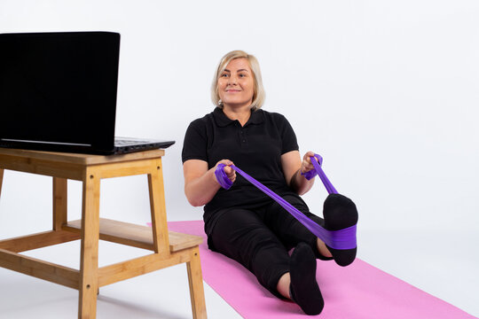 An Older Woman Sits On A Mat In Front Of A Computer And Does Exercises With An Elastic Band. Pandemic And Senior Online Concept. Photo On White Background