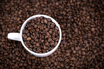White cup full of coffee beans close up against the coffee beans background. Coffee mug. Morning espresso.