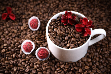 White cup full of coffee beans on a coffee beans background with red chocolate candy and red flowers. Morning espresso. Coffee mug.