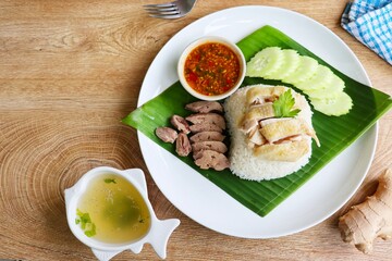 Hainanese chicken rice,Thai gormet steamed chicken with rice served with steamed chicken livers,sliced cucumber,sauce and hot soup in bowl on banana leaf and white plate with wooden table background