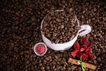 White cup full of coffee beans on a coffee beans background with red flowers , red chocolate and cinnamon. Morning espresso. Coffee mug.