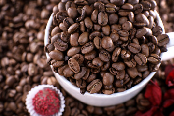 White cup full of coffee beans on a coffee beans background with red chocolate candy and red flowers. Morning espresso. Coffee mug.