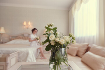 A girl in a white negligee is sitting on the bed with a bouquet of white roses in her hand