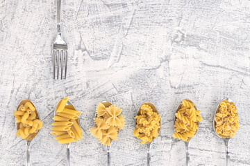 One fork and various pasta on spoons over white stone background. Top view copy space