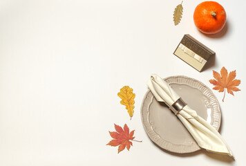 A ringed napkin on a gray plate and an empty name card on a white table with a small orange pumpkin and bright autumn leaves