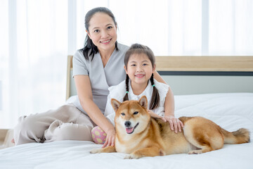 Asian mother and girl sit on white bed with Shiba dog and look to camera with smiling in bedroom. Concept of happy family feel relax to stay home with their own pets.