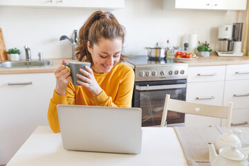 Mobile Office at home. Young woman sitting in kitchen at home working using on laptop computer. Lifestyle girl studying or working indoors. Freelance business quarantine concept.