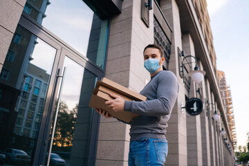 Low-angle shot of delivery man wearing medical mask ringing door for delivery carton boxes with hot pizza, looking at camera. Deliveryman in protective mask holding box with food.