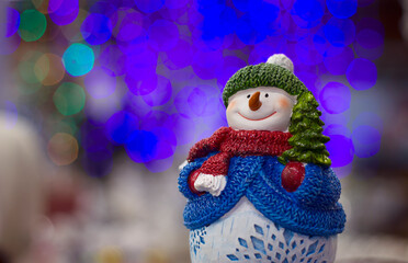 Happy smiling snowman in knitted hat and clothes, with a little green spruce tree under the arm, with unfocused and blurred colorful lights background, merry christmas and happy new year, banner, card
