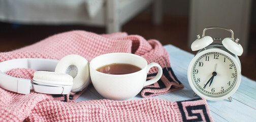 A cup of hot tea, knitted sweater, alarm clock and headphones in the bedroom. winter morning concept. banner