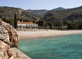 Rocks, blue sea coming onto the gravel beach, mountains and a two storey building. Queen's Beach, Montenegro.