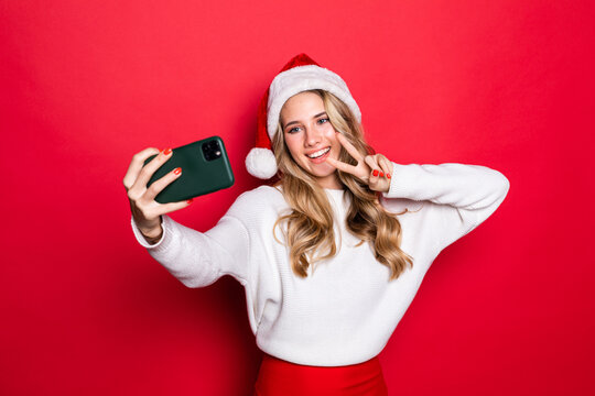 Portrait of a pretty woman with peace gesture wearing Christmas hat taking a selfie isolated over red background