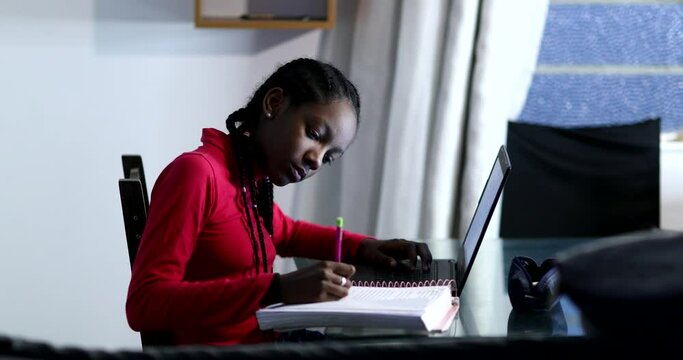 Teen African girl doing homework at night, black adolescent female writing note in front of computer