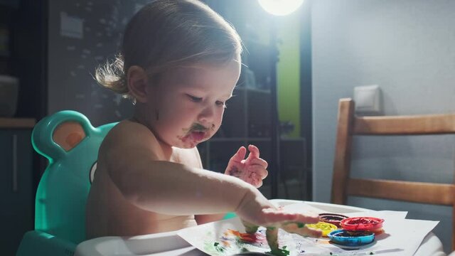 Baby paints. Infant baby with paint stained face plays with water paints in the room