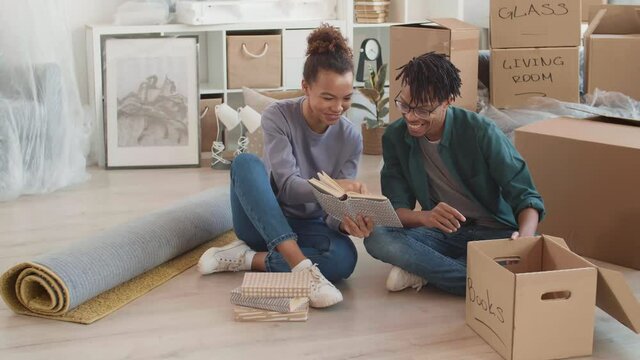 Wide shot of young multiethnic couple sitting on floor indoors and looking through books while packing things for move
