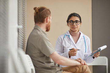 Young female doctor holding medical card and discussing with patient his disease while they sitting...