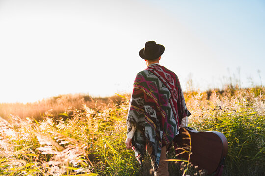 Man with acoustic guitar wearing poncho in a rural area at sunset