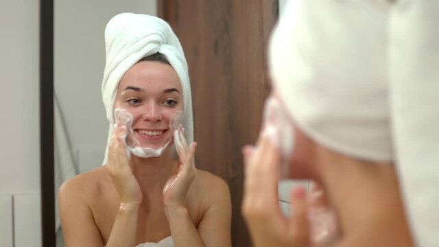 Young cute attractive smiling woman wearing white bath towel looking at herself at bathroom mirror and washing face with facial foam after shower