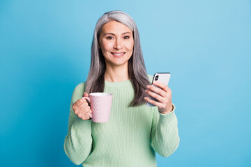 Photo of retired old woman arm hold cup smartphone look camera wear green sweater isolated blue color background