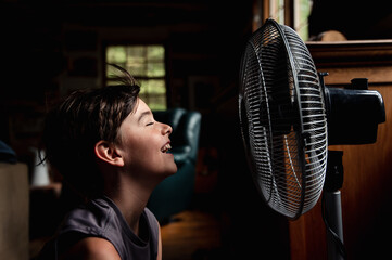 Young boy with eyes closed cooling off in front of a fan in dark room.