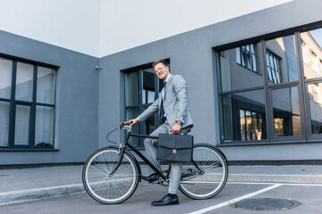 Businessman smiling at camera while holding briefcase and riding bike outdoors