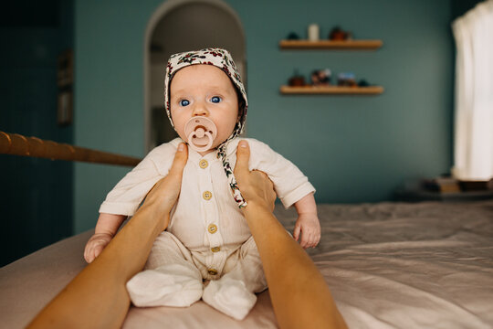 Young infant in a bonnet sitting on bed with help from a mothers hands