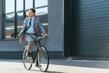 Fototapeta premium Positive businessman in formal wear and eyeglasses riding bicycle near building