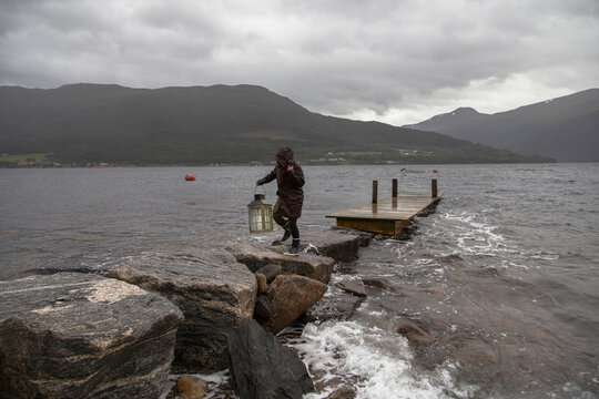 A woman carrying a lantern along a stone jetty next to a fjord