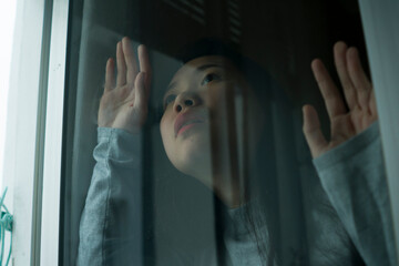 young sad and depressed Asian Chinese girl scared and worried during covid19 lockdown and stay home quarantine looking through window feeling unhappy and stressed