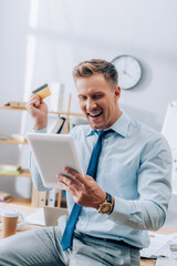 Obraz premium Cheerful businessman looking at digital tablet while holding credit card