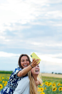 A couple of attractive young women one blonde and the other brunette posing in their designer dresses in a field of sunflowers using their smartphone