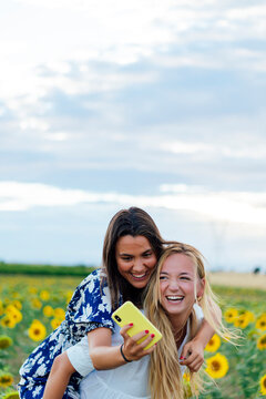 A couple of attractive young women one blonde and the other brunette posing in their designer dresses in a field of sunflowers using their smartphone