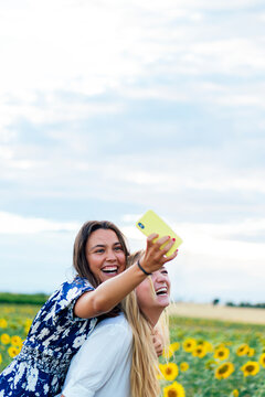 A couple of attractive young women one blonde and the other brunette posing in their designer dresses in a field of sunflowers using their smartphone