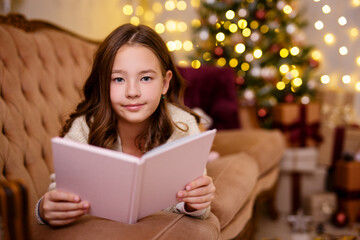 Christmas concept - little girl reading book in decorated living room with Christmas tree and lights